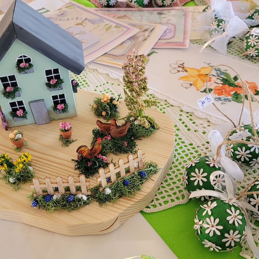 Eine Miniatur-Gartenszene mit einem Haus, Blumen, einem Hahn und Ostereiern auf einem Holzbrett, mit einem Spitzentischtuch im Hintergrund.