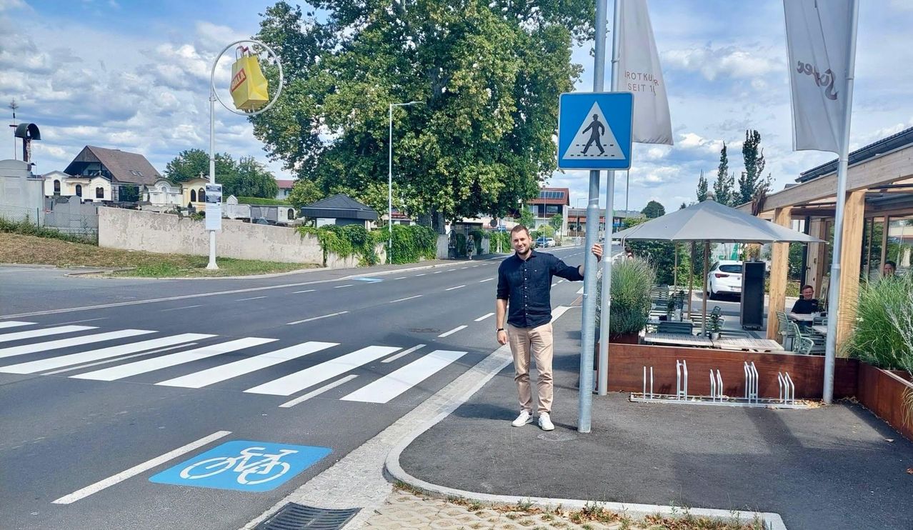 A man stands by a pole on a street with a pedestrian crossing. A yellow bag hangs above him, and a bicycle symbol is painted on the pavement.