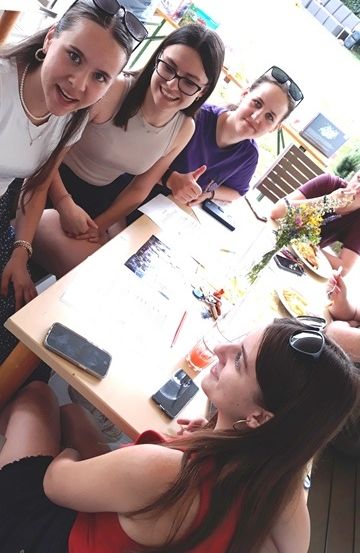 Eine Gruppe von Frauen sitzt um einen Tisch herum, lächelt und posiert für ein Foto. Sie befinden sich in einem Outdoor-Restaurant mit Stühlen und Tischen. Auf dem Tisch sind Teller, Blumen und ein Handy.