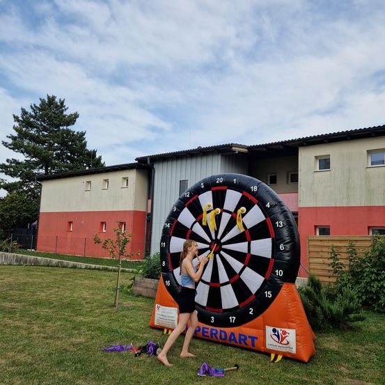 Ein junges Mädchen spielt Darts auf einem großen aufblasbaren Dartboard in einer Rasenfläche vor einem Gebäude.