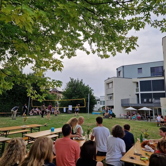 Eine Outdoor-Veranstaltung mit einem Volleyballnetz in der Mitte eines Grasfeldes. Menschen sitzen auf Bänken und Tischen, während andere Volleyball spielen. Im Hintergrund befinden sich Gebäude mit einigen Menschen, die draußen sitzen.