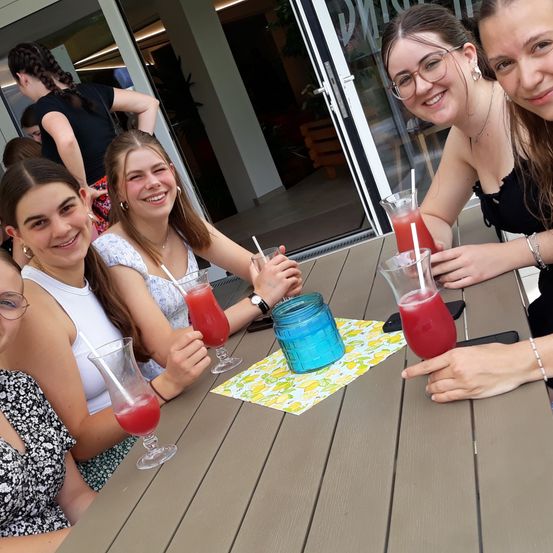 Fünf Mädchen sitzen um einen Tisch herum, lächeln und halten rote Getränke in hohen Gläsern. Sie scheinen für ein Foto zu posieren.