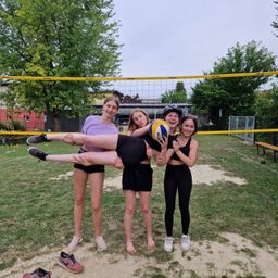 Vier Mädchen stehen lächelnd in der Nähe eines Volleyballnetzes. Ein Mädchen hält einen Volleyball, während ein anderes mit erhobenen Beinen auf dem Boden liegt.