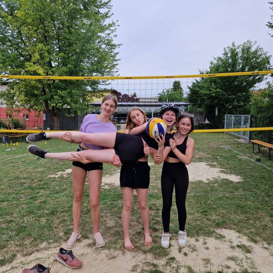 Vier Mädchen stehen lächelnd in der Nähe eines Volleyballnetzes. Ein Mädchen hält einen Volleyball, während ein anderes mit erhobenen Beinen auf dem Boden liegt.