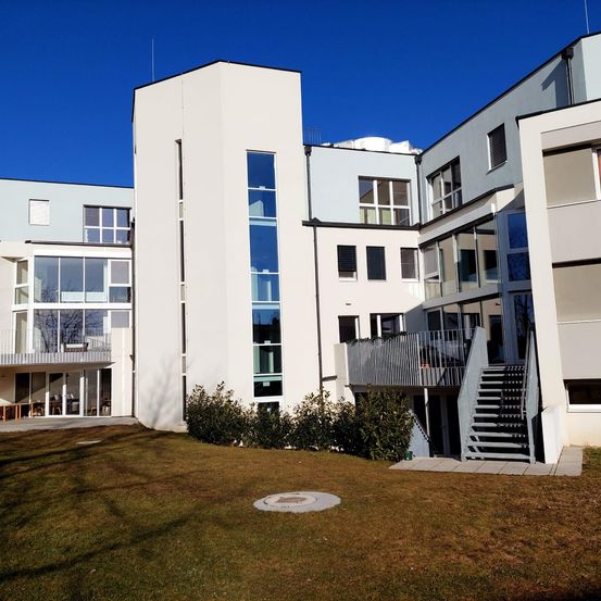 Ein modernes weißes Gebäude mit mehreren Stockwerken, großen Glasfenstern und einer Treppe, die zu einem Balkon führt, vor einem klaren blauen Himmel.