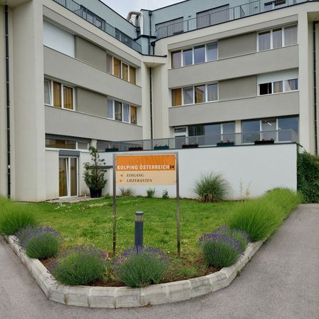 Vogelperspektive eines mehrstöckigen Gebäudes mit mehreren Fenstern und einem Schild, das 'Kolping Osterreich' steht. Das Gebäude ist von einem Rasen mit Lavendelpflanzen umgeben.