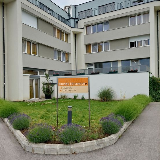 Vogelperspektive eines mehrstöckigen Gebäudes mit mehreren Fenstern und einem Schild, das 'Kolping Osterreich' steht. Das Gebäude ist von einem Rasen mit Lavendelpflanzen umgeben.