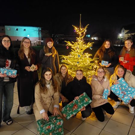 Eine Gruppe von Frauen posiert nachts für ein Foto, alle halten Geschenke in der Hand, mit einem geschmückten Weihnachtsbaum im Hintergrund.