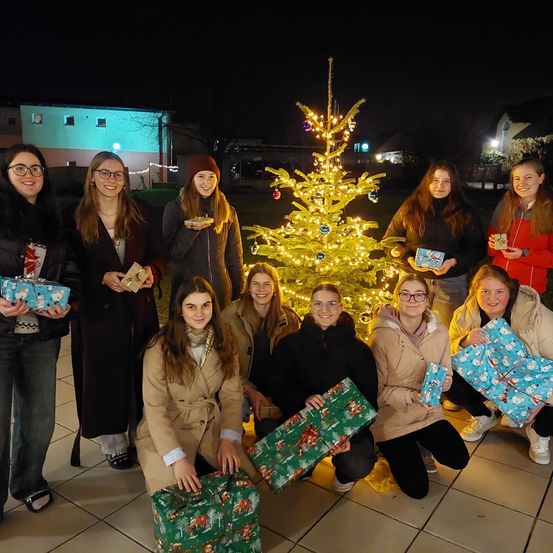 Eine Gruppe von Frauen posiert nachts für ein Foto, alle halten Geschenke in der Hand, mit einem geschmückten Weihnachtsbaum im Hintergrund.