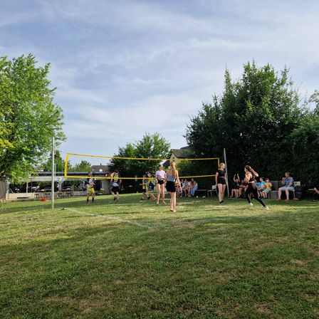 Eine Gruppe von Menschen spielt Volleyball auf einem Rasenfeld unter einem klaren Himmel. Einige rennen und andere stehen still.
