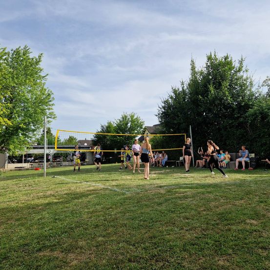 Eine Gruppe von Menschen spielt Volleyball auf einem Rasenfeld unter einem klaren Himmel. Einige rennen und andere stehen still.