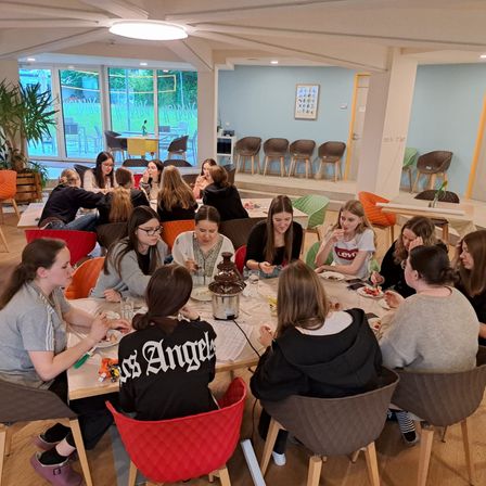Eine Gruppe von Frauen sitzt um einen Tisch in einem Raum, isst und plaudert. Eine Schokoladenfontäne steht auf dem Tisch. Der Raum hat Holzböden, eine Deckenlampe und große Fenster.