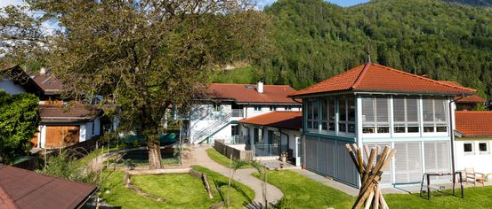 Luftaufnahme eines kleinen Dorfes mit einem großen Baum und rot gedeckten Häusern vor dem Hintergrund grüner Hügel.