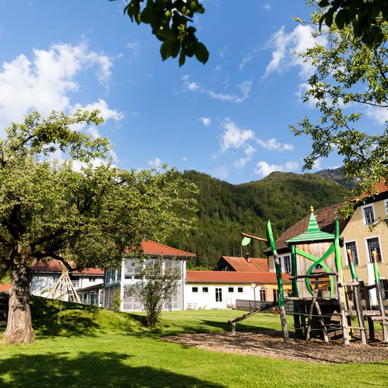 Ein sonniger Tagesausblick auf einen Rasenspielplatz mit einem Baum und einem Berg im Hintergrund. Ein grünes Spielhaus befindet sich rechts.
