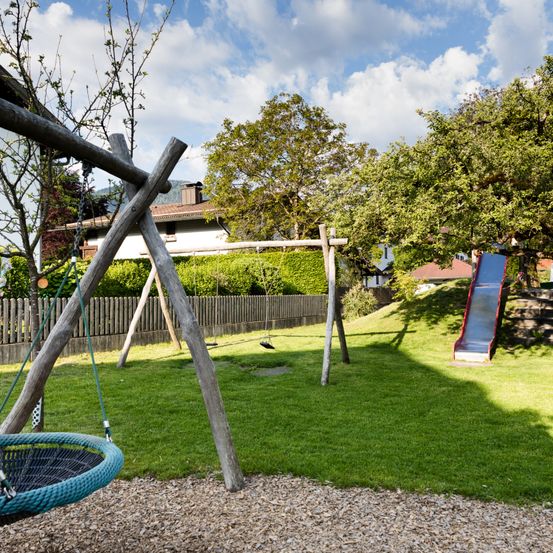 Ein Kinderspielplatz mit Schaukel, Rutsche und zwei Schaukeln, in einem Rasenbereich mit Holzzaun. Hinter dem Zaun ist ein Haus mit Schornstein sichtbar.