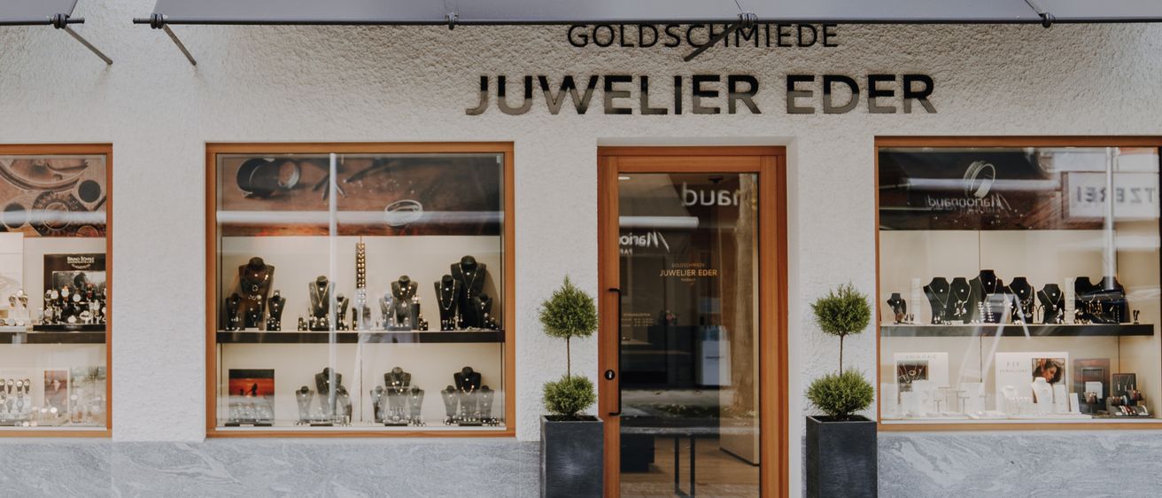 Ein Juweliergeschäft mit dem Namen Goldsco Juwelier. Zwei Glasvitrinen zeigen verschiedene Schmuckstücke, neben der Tür steht ein Topf mit einer Pflanze.