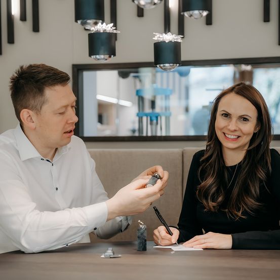 Ein Mann und eine Frau sitzen an einem Tisch, der Mann zeigt der Frau etwas, die lächelt und auf einem Blatt Papier schreibt.