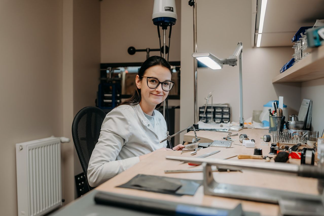Eine Frau in einem Laborkittel sitzt lächelnd am Schreibtisch. Hinter ihr befinden sich Laborgeräte und ein Mikroskop. Eine Lampe und ein Heizkörper sind an der Seite.