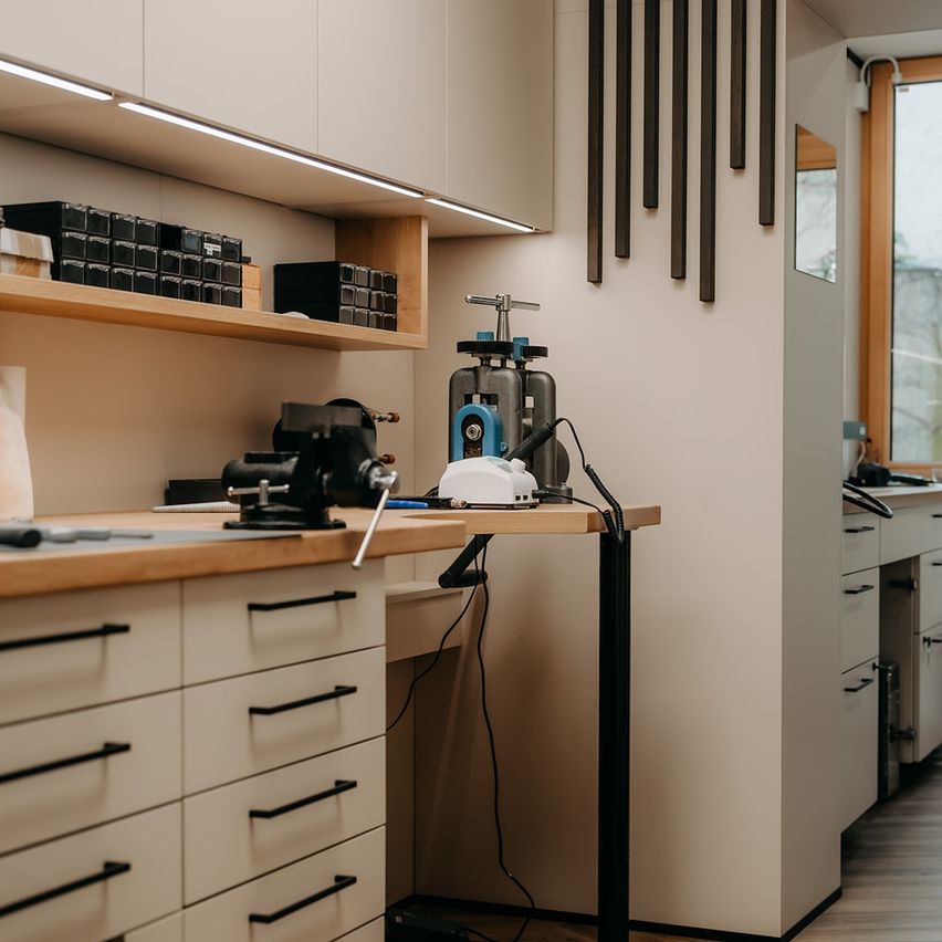 Ein Werkstattraum mit einem Metallarbeitstisch, Werkzeugen und Regalen. Eine Glastür führt in einen anderen Raum.