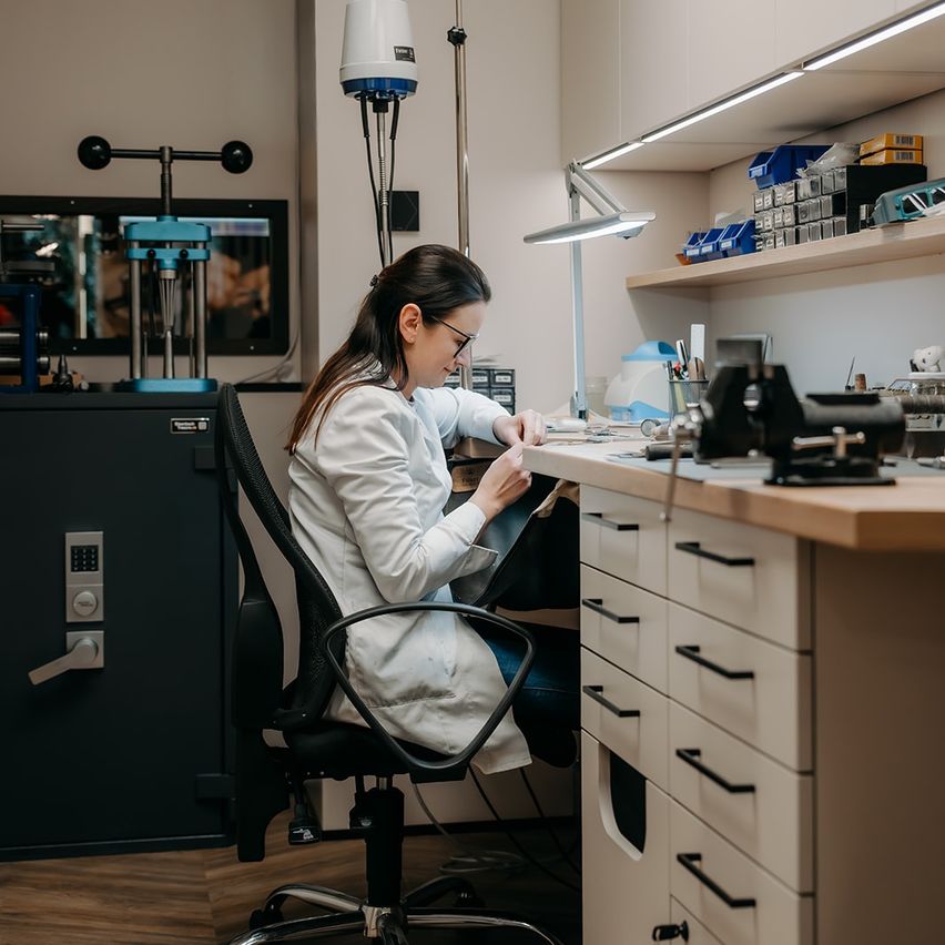 Eine Frau in einem weißen Laborkittel arbeitet an einem Schreibtisch in einem Labor, umgeben von verschiedenen Werkzeugen und Geräten.