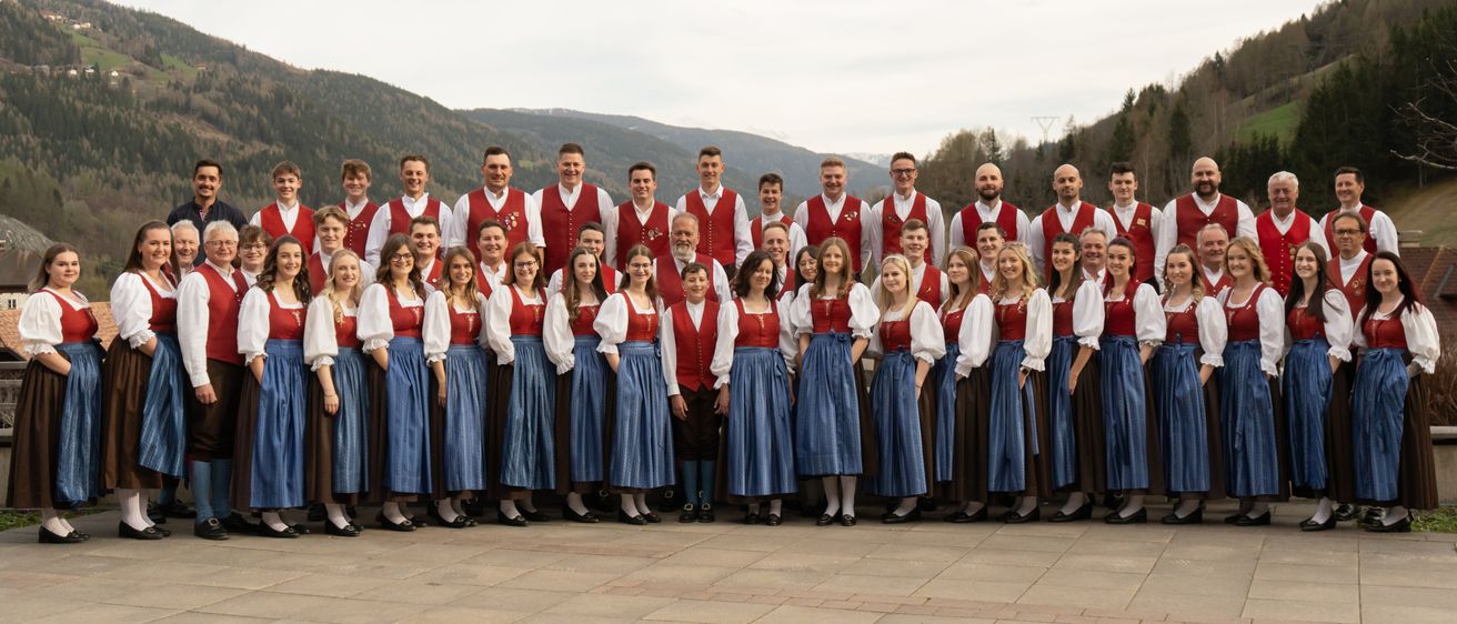 Ein Chorensemble in traditioneller deutscher Kleidung posiert für ein Foto vor einem Berg.