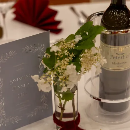 Eine Tischdekoration mit einer Flasche Peterhof-Wein, einer Glasvase mit weißen Blumen und einer Karte mit der Aufschrift 'Swinging Dinner 2023'. Ein rotes Serviettentuch und Besteck sind ebenfalls vorhanden.