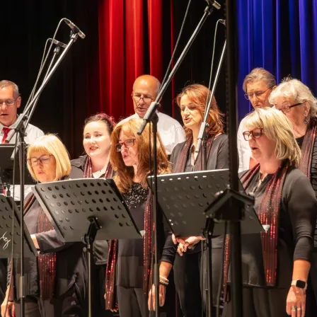 Ein Chor von Frauen tritt auf der Bühne auf, mit Mikrofonen vor ihnen. Sie sind in Schwarz gekleidet und haben rote Schals. Hinter ihnen steht ein Mann mit Krawatte und eine Frau mit Brille. Die Bühne hat blaue Vorhänge und der Hintergrund ist dunkel.