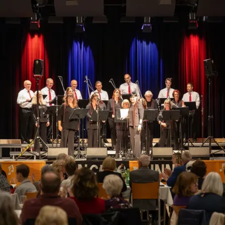 Ein Chor tritt auf der Bühne mit einem blau-roten Vorhang im Hintergrund auf. Sie werden von einem Pianisten begleitet, und das Publikum sitzt vor ihnen.