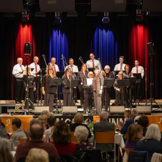 Ein Chor tritt auf der Bühne mit einem blau-roten Vorhang im Hintergrund auf. Sie werden von einem Pianisten begleitet, und das Publikum sitzt vor ihnen.