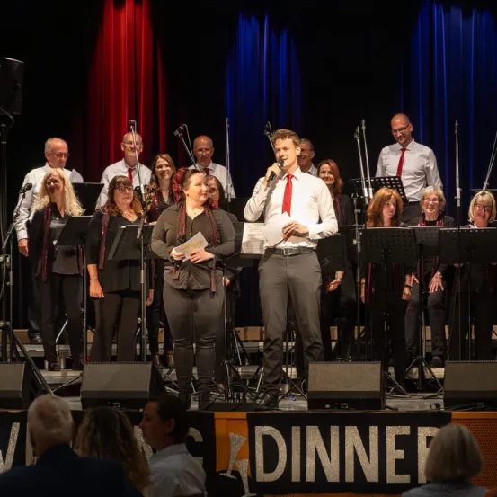 Ein Chor tritt auf der Bühne auf, wobei ein Mann ein Mikrofon hält und singt. Sie sind von Notenständern und Lautsprechern umgeben. Die Bühne ist mit blauen Vorhängen dekoriert.