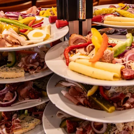 Ein Tisch ist mit mehreren weißen Tellern gefüllt, die aufeinander gestapelt sind, jeder mit einer Auswahl an Lebensmitteln. Der oberste Teller hat geschnittenes Fleisch, Eier und Gemüse. Der mittlere Teller hat mehr geschnittenes Fleisch, Gemüse und Käse. Der unterste Teller hat mehr Fleisch, Zwiebeln und Gurken.