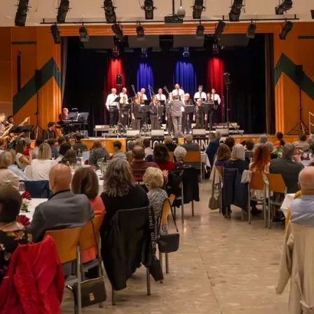Eine Band tritt auf einer Bühne vor einem Publikum auf, das an Tischen sitzt, einige mit Weingläsern. Die Bühne ist mit blauen und roten Vorhängen geschmückt.