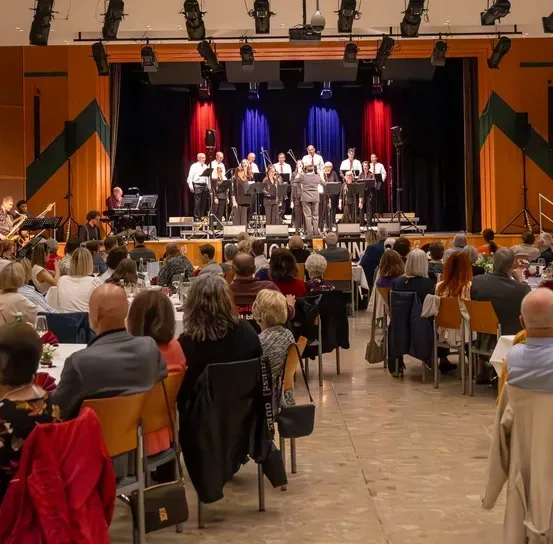 Eine Band tritt auf einer Bühne vor einem Publikum auf, das an Tischen sitzt, einige mit Weingläsern. Die Bühne ist mit blauen und roten Vorhängen geschmückt.