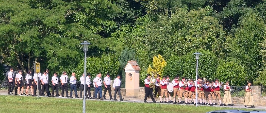 Eine Gruppe von Personen in traditionellen Uniformen marschiert mit Musikinstrumenten. Sie gehen auf einem Weg, der von Bäumen und Gras umgeben ist. Rechts sind drei geparkte Autos.