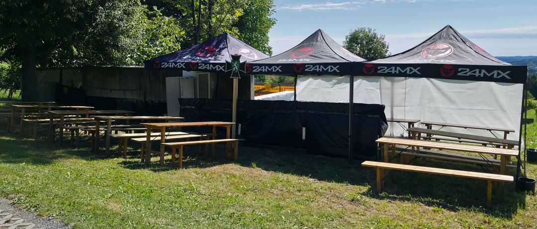 Drei schwarze Zelte mit dem 24MX-Logo sind in einem Grasbereich aufgestellt. Holztische und Bänke sind um die Zelte herum angeordnet, mit einem weiteren Zelt, das teilweise auf der rechten Seite sichtbar ist. Bäume und ein klarer Himmel sind im Hintergrund zu sehen.