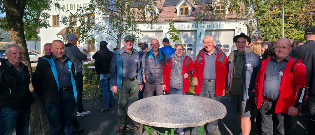 Eine Gruppe von Menschen steht vor einem Gebäude, lächelt und posiert für ein Foto. Das Gebäude hat Solarpanele auf dem Dach und ist als 'Feuerwehr' beschriftet. Sie sind für kühleres Wetter gekleidet.