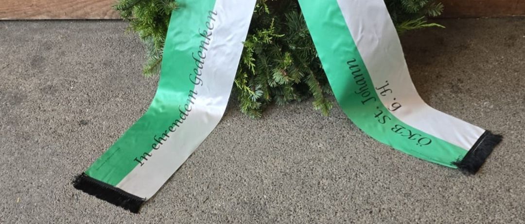 Ein Kranz mit grünen und weißen Bändern und einer Schleife, auf einem Betonboden vor einer Holzwand platziert. Das Band trägt die Aufschrift 'In Ehrenden Gedenken'.