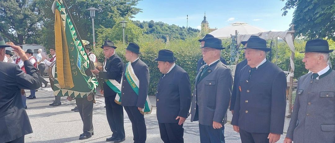 Fünf Männer in formeller Kleidung stehen in einer Reihe, möglicherweise bei einer Parade. Ein Mann hält eine Fahne. Bäume und ein weißes Zelt sind im Hintergrund.