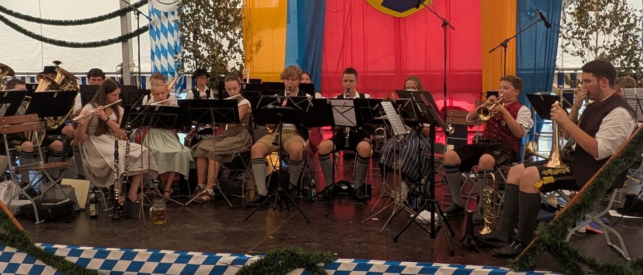 Ein Blasorchester spielt auf einer Bühne unter einem Zelt mit einem Banner, das 'FV Oberaudorf 1961' steht. Die Bühne ist mit blau-weiß kariertem Stoff dekoriert.