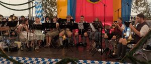 Ein Blasorchester spielt auf einer Bühne unter einem Zelt mit einem Banner, das 'FV Oberaudorf 1961' steht. Die Bühne ist mit blau-weiß kariertem Stoff dekoriert.