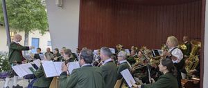 Eine Blaskapelle sitzt in einer kreisförmigen Formation und spielt verschiedene Instrumente. Sie befinden sich in einem Raum mit Holzwänden und Deckenleuchten. Einige Musiker tragen traditionelle Kleidung.