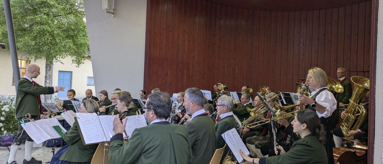 Eine Blaskapelle sitzt in einer kreisförmigen Formation und spielt verschiedene Instrumente. Sie befinden sich in einem Raum mit Holzwänden und Deckenleuchten. Einige Musiker tragen traditionelle Kleidung.