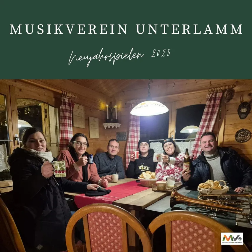 Eine Gruppe von Menschen sitzt in einer Hütte um einen Tisch herum, lächelt und hält Tassen. Sie scheinen eine Mahlzeit gemeinsam zu genießen. Der Tisch ist mit Brot, Tassen und einer Flasche gedeckt. Eine Trompete steht auf dem Tisch. Der Raum ist warm beleuchtet, eine Lampe hängt von der Decke. Das Bild hat einen Wasserzeichen in der unteren rechten Ecke.