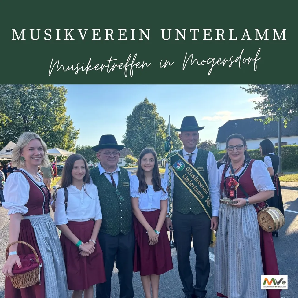 Eine Gruppe von Menschen in traditioneller deutscher Kleidung, die vor einem Gebäude mit einem Banner steht, auf dem 'Musikverein Unterlamm Musiktreffen in Mogersdorf' steht. Das Bild fängt einen Moment der Feier mit einer festlichen Atmosphäre ein.