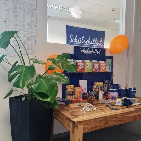 Ein Holztisch zeigt eine Vielzahl bunter Boxen, eine Pflanze und Luftballons. Ein Banner mit dem Text 'Schülerhilfe' befindet sich über dem Tisch.