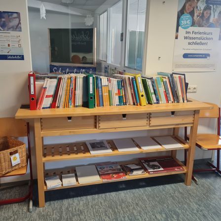 Ein Holzschreibtisch mit Schubladen und Büchern, mit einem Korb auf einem Stuhl, an einer Wand mit einem Poster. Es gibt eine Reflexion eines Klassenzimmers und eines weiteren Posters auf der Glasscheibe.