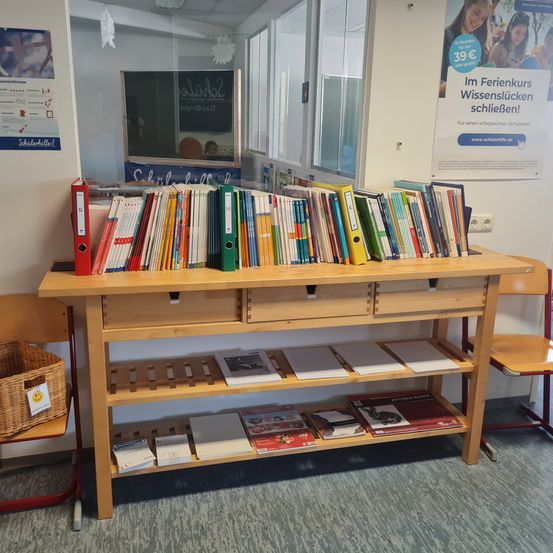 Ein Holzschreibtisch mit Schubladen und Büchern, mit einem Korb auf einem Stuhl, an einer Wand mit einem Poster. Es gibt eine Reflexion eines Klassenzimmers und eines weiteren Posters auf der Glasscheibe.