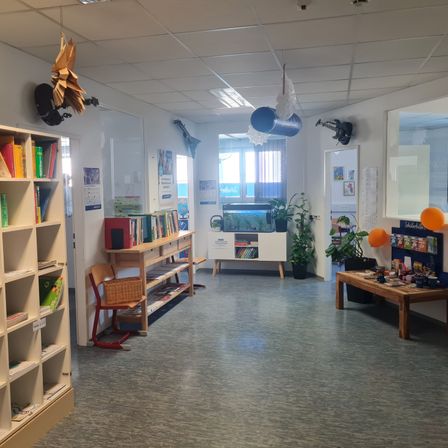 Ein leerer Raum hat Bücherregale, Stühle, einen Tisch, Pflanzen und Luftballons. Eine Glastür führt in einen Raum mit einem Aquarium und Aquarium.