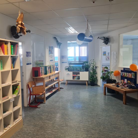 Ein leerer Raum hat Bücherregale, Stühle, einen Tisch, Pflanzen und Luftballons. Eine Glastür führt in einen Raum mit einem Aquarium und Aquarium.