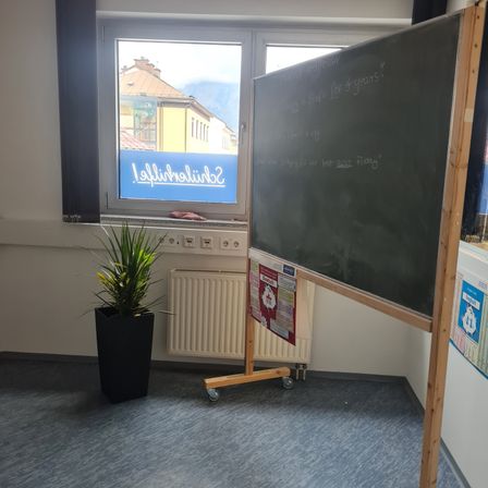 Ein leeres Klassenzimmer mit einer großen Tafel mit Schrift, einem Topfpflanzen und einem Fenster mit einem blauen Schild.